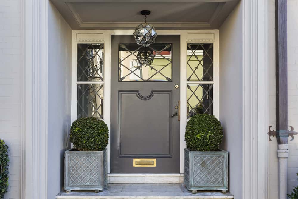 A gray front door with decorative glass panels, flanked by two potted round bushes, and a geometric hanging light above