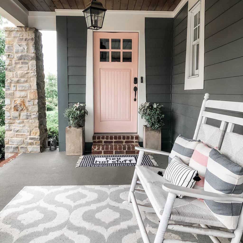 Cozy porch with a pastel pink door, white rocking chair, patterned pillows, and potted plants on either side of the entrance