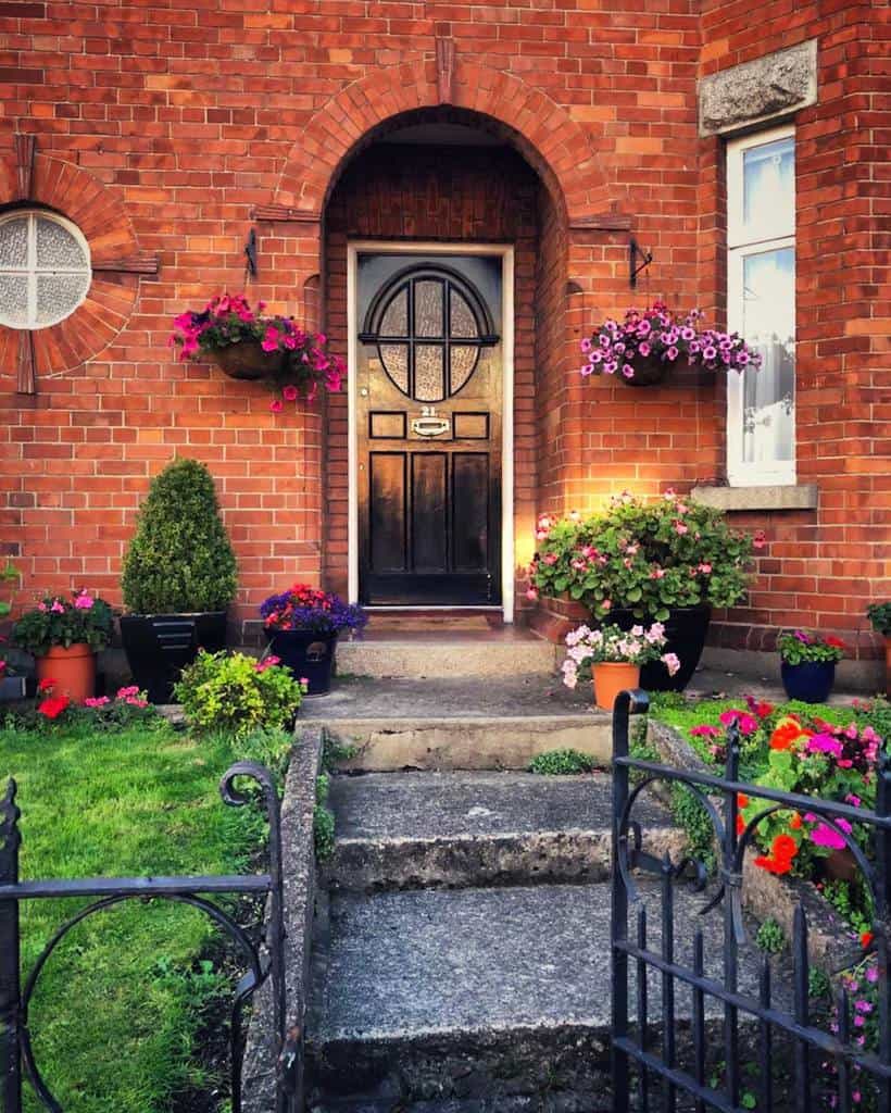 A brick house entrance with an arched black door, surrounded by colorful flowers and potted plants on either side of the steps