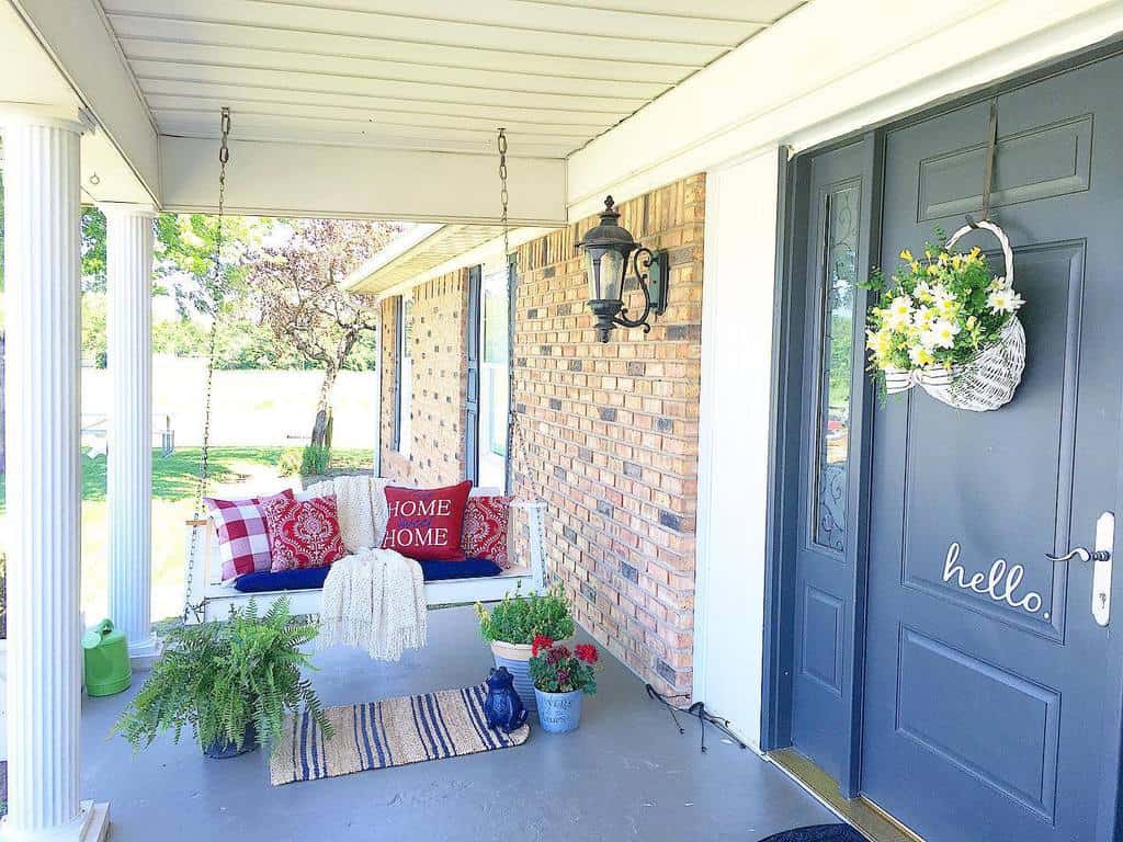 Front porch with a swinging bench, red and white pillows, potted plants, and a dark blue door with "hello" written on it