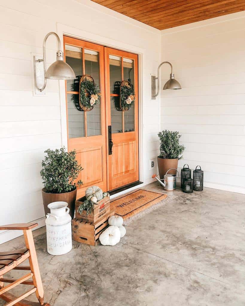 Inviting porch with wooden double doors, wreaths, potted plants, lanterns, pumpkins, and a vintage milk can beside a rocking chair