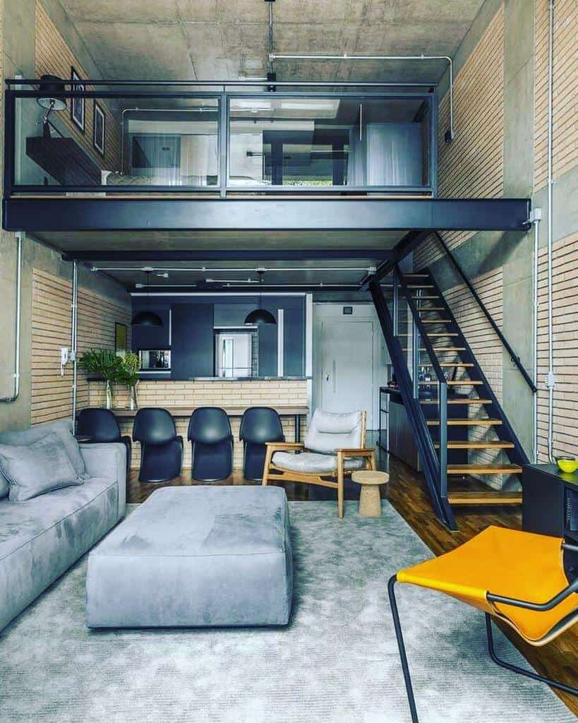 A modern loft with a gray sofa, black chairs, and a yellow chair, featuring a mezzanine with a bed overlooking the open kitchen and living area.