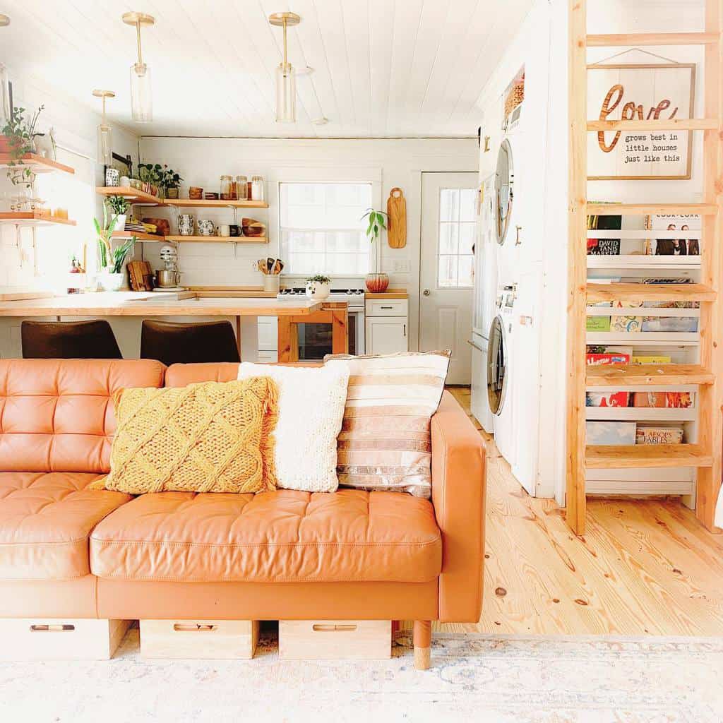 Cozy living room with a brown sofa, cushions, and open kitchen; wooden ladder leads to loft; light, airy decor with shelves and plants.