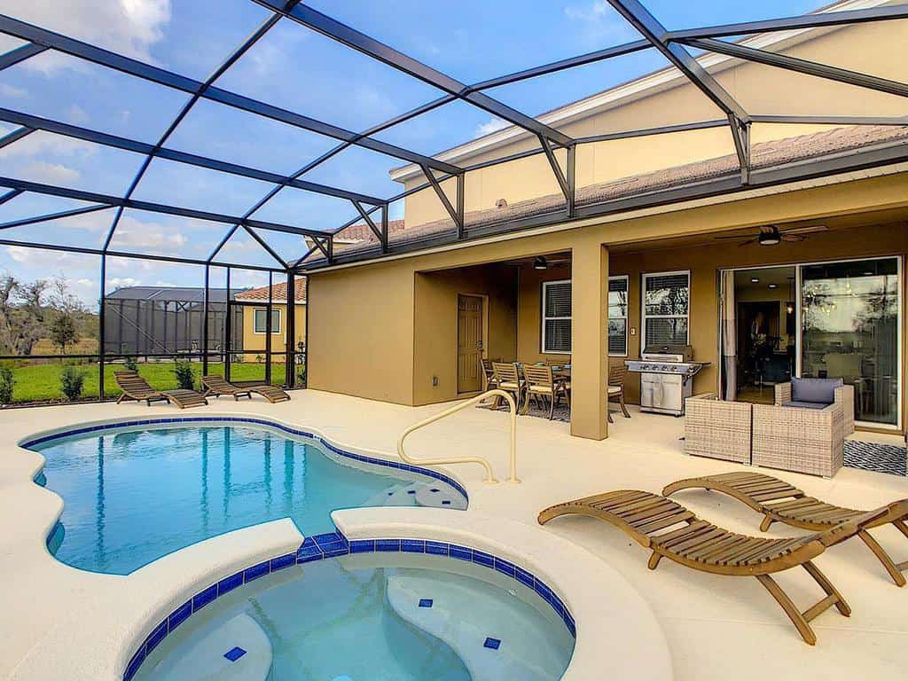 Outdoor pool area with a jacuzzi, lounge chairs, dining table, and covered patio surrounded by a screened enclosure under a blue sky