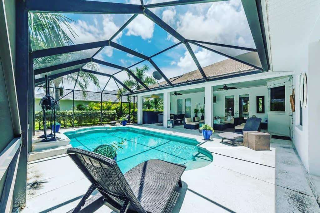 Sunny patio with a covered pool, lounge chairs, and seating area surrounded by palm trees and greenery under a blue sky