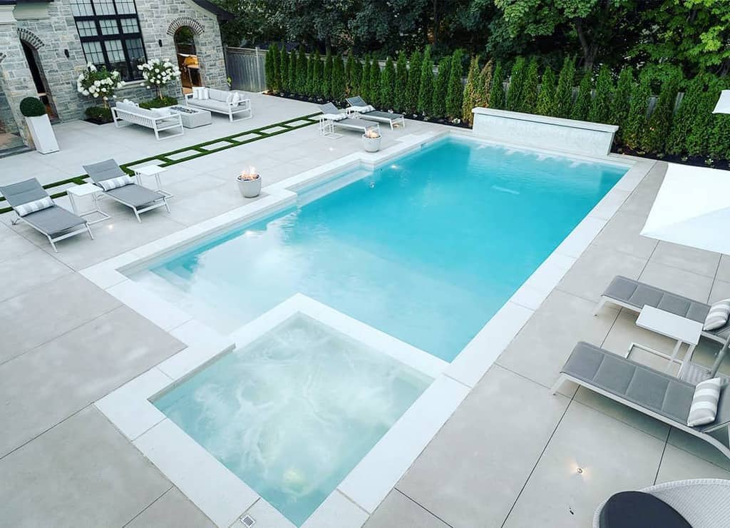 Modern backyard with a rectangular pool, adjacent hot tub, lounge chairs, and greenery along the fence