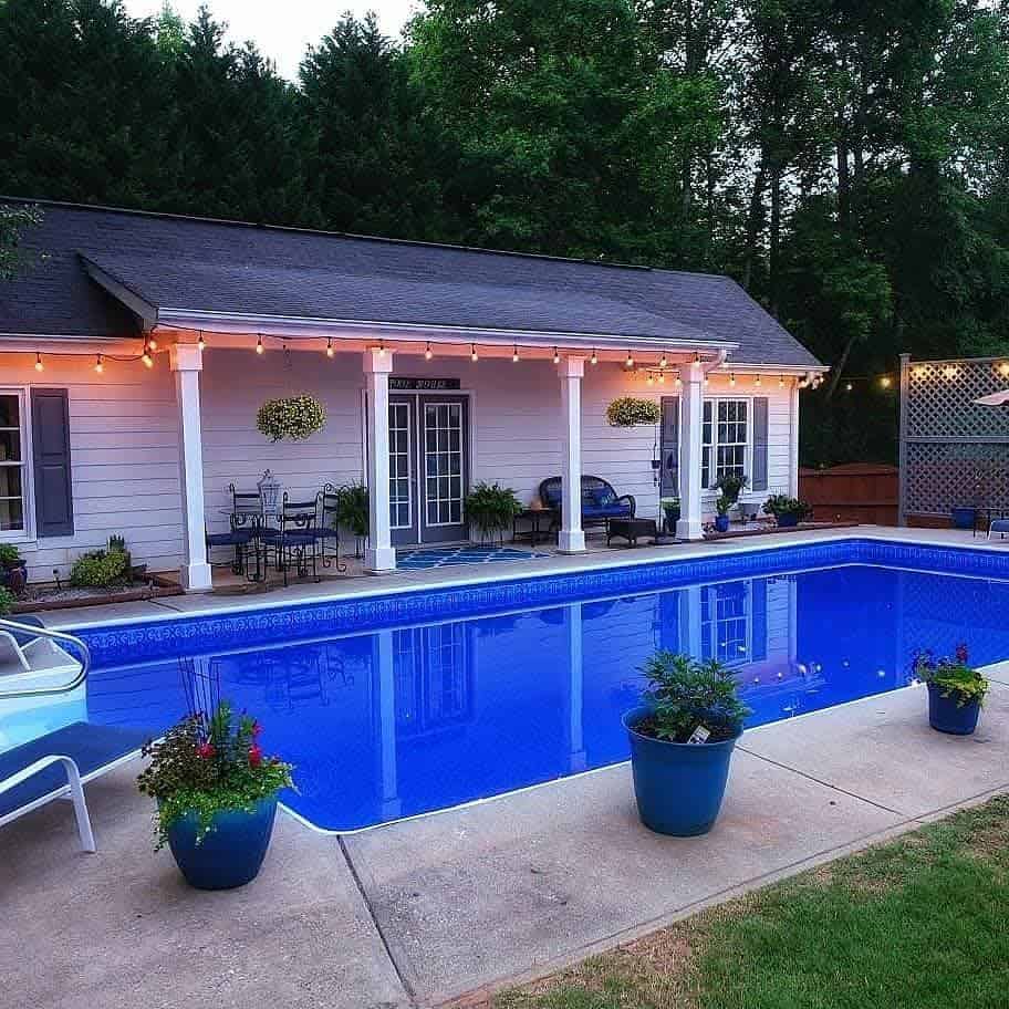 Cozy backyard with a pool, lighted pool house, and potted plants. White loungers and chairs create a relaxing atmosphere