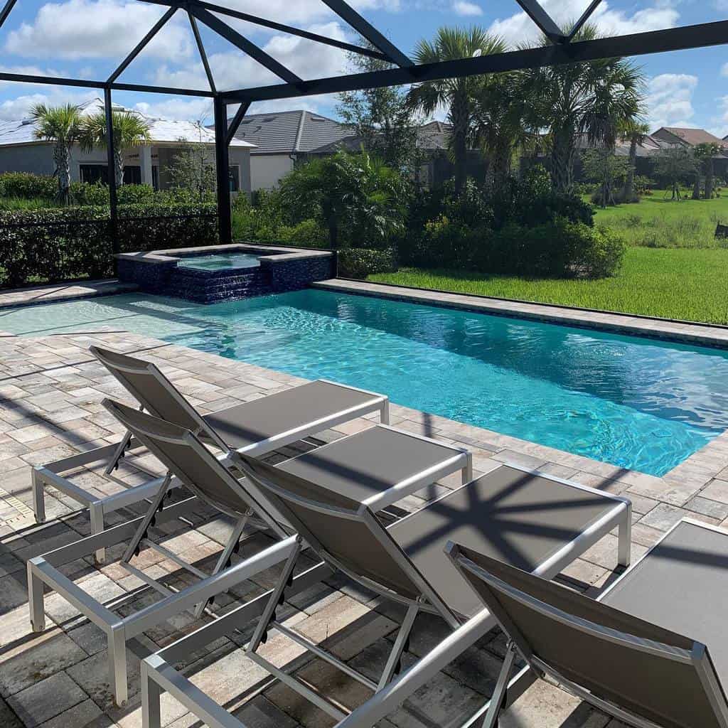 Outdoor pool area with four gray lounge chairs on a stone-paved deck, a hot tub, and a view of grassy landscape and houses behind