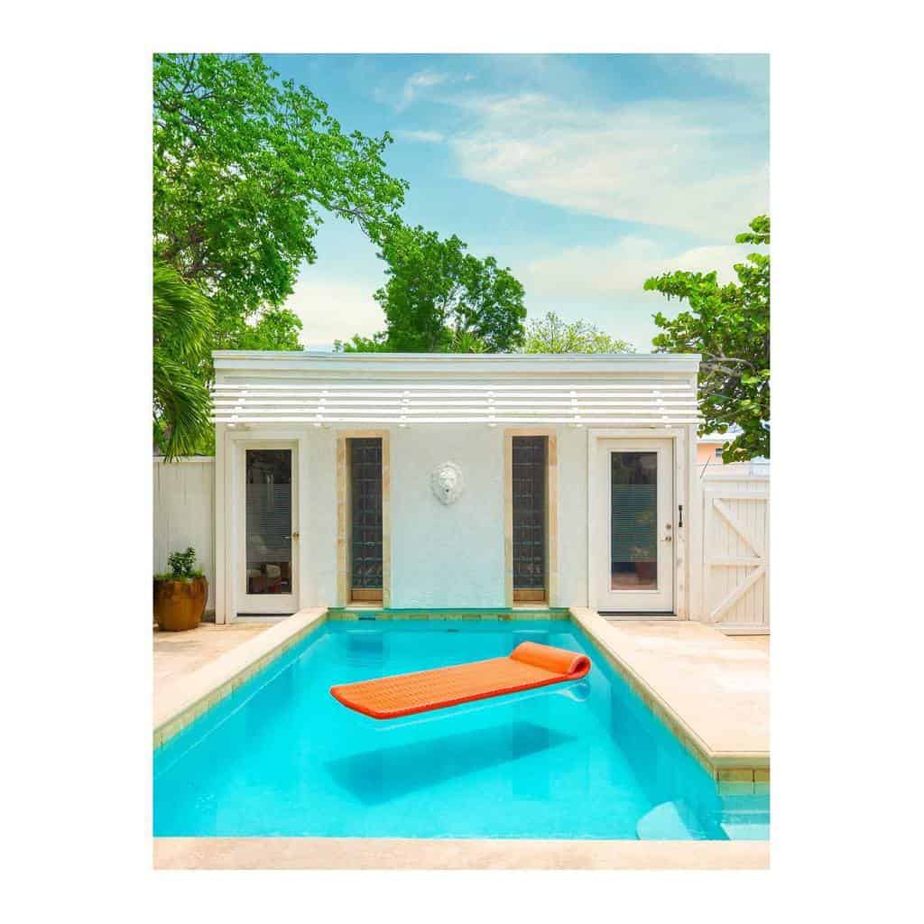 A bright orange float lies on a rectangular pool beside a modern white building, surrounded by lush green trees