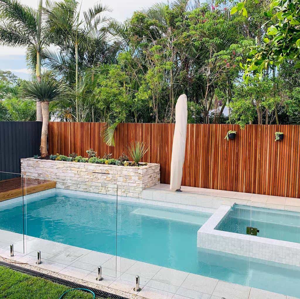 Modern backyard with a glass-fenced pool, surrounded by palm trees and plants, and a stone planter against a wooden fence