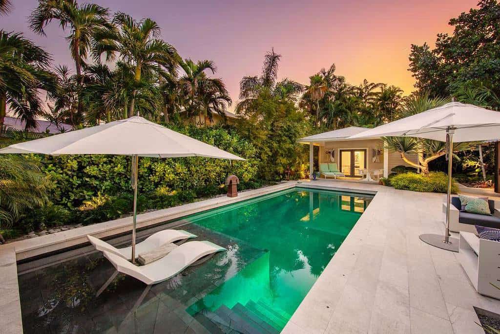 Tropical backyard with a pool, two lounge chairs, and white umbrellas set amid lush greenery and a sunset sky in the background