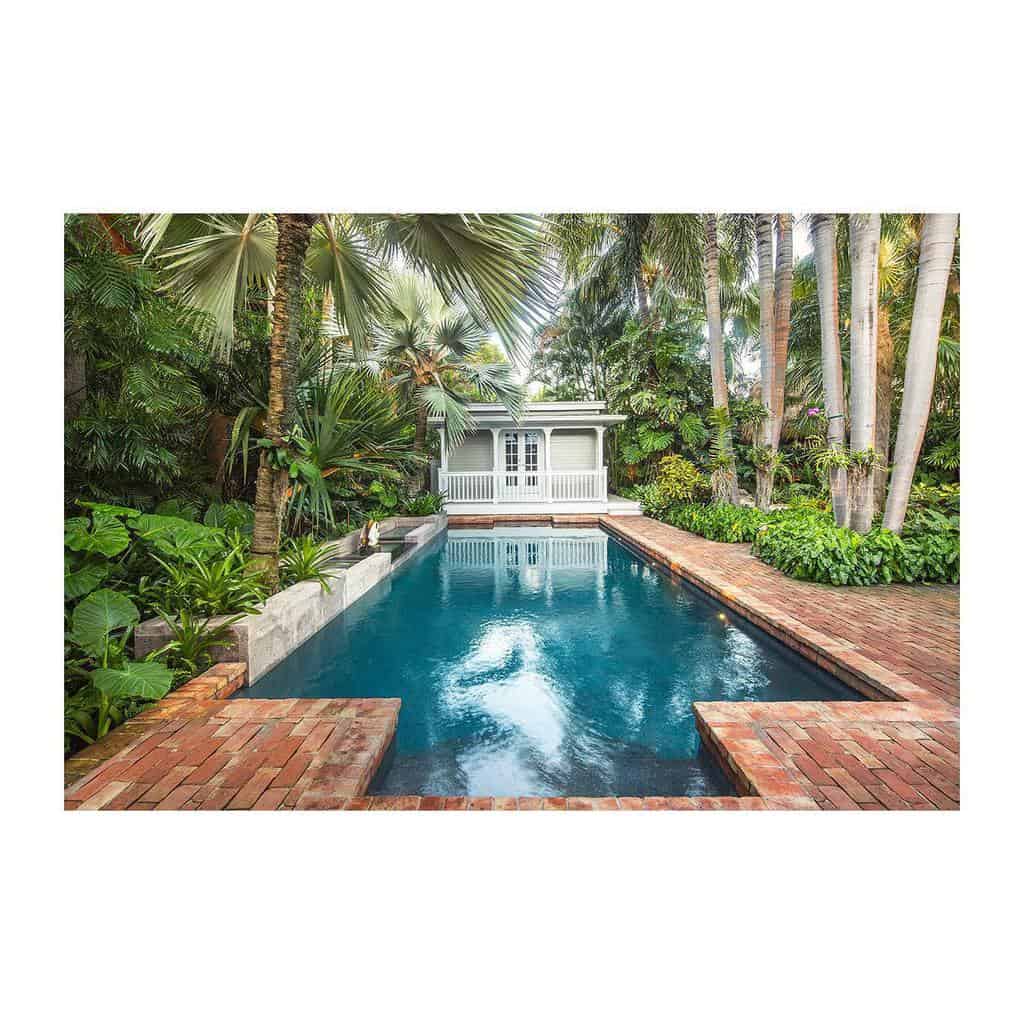 A tropical backyard with a rectangular pool, surrounded by lush greenery and brick paths, leading to a small white villa