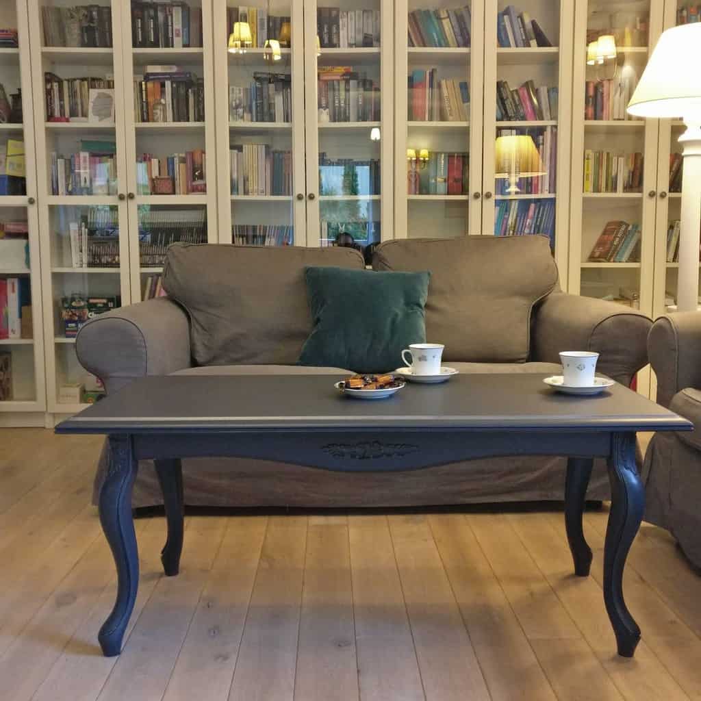 Elegant dark-painted wooden coffee table with curved legs, set in a cozy reading nook with bookshelves, a plush sofa, and warm lighting