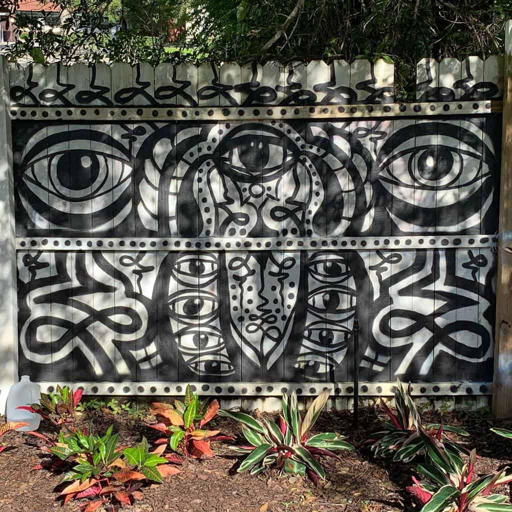 Bold black and white mural on a wooden fence featuring abstract eyes and patterns, creating a striking artistic statement in the garden