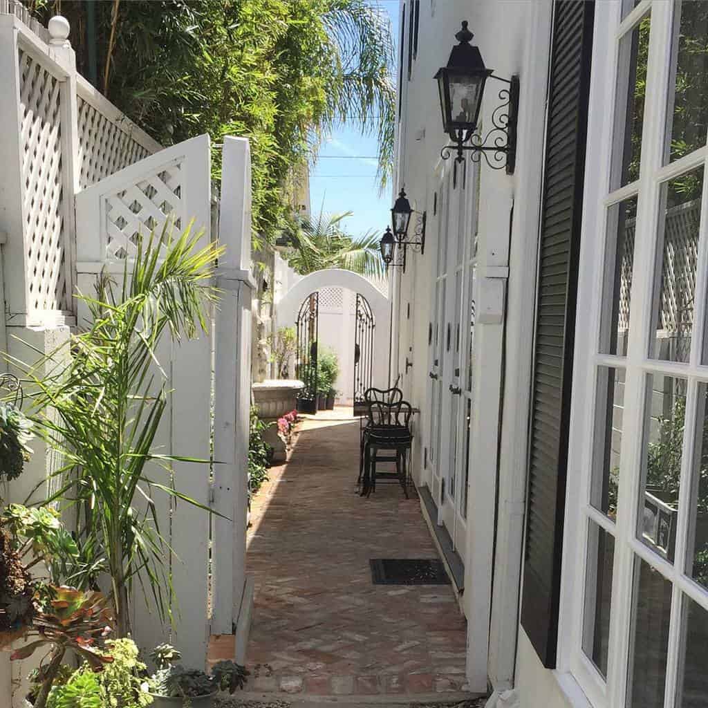 Narrow outdoor brick pathway with plants, white lattice fence, a black wrought iron chair and lanterns, leading to an arched white gate