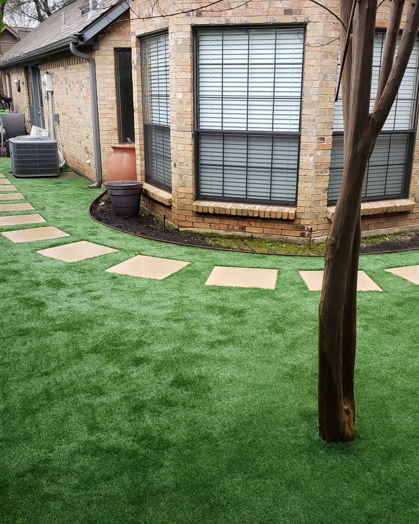 Side yard with artificial grass, a curved stepping stone path, a brick house with large windows, potted plants, and a tree