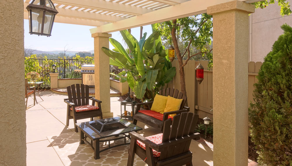 Outdoor patio with wooden chairs and cushions around a glass table, under a pergola; lush plants and a barbecue grill in the background