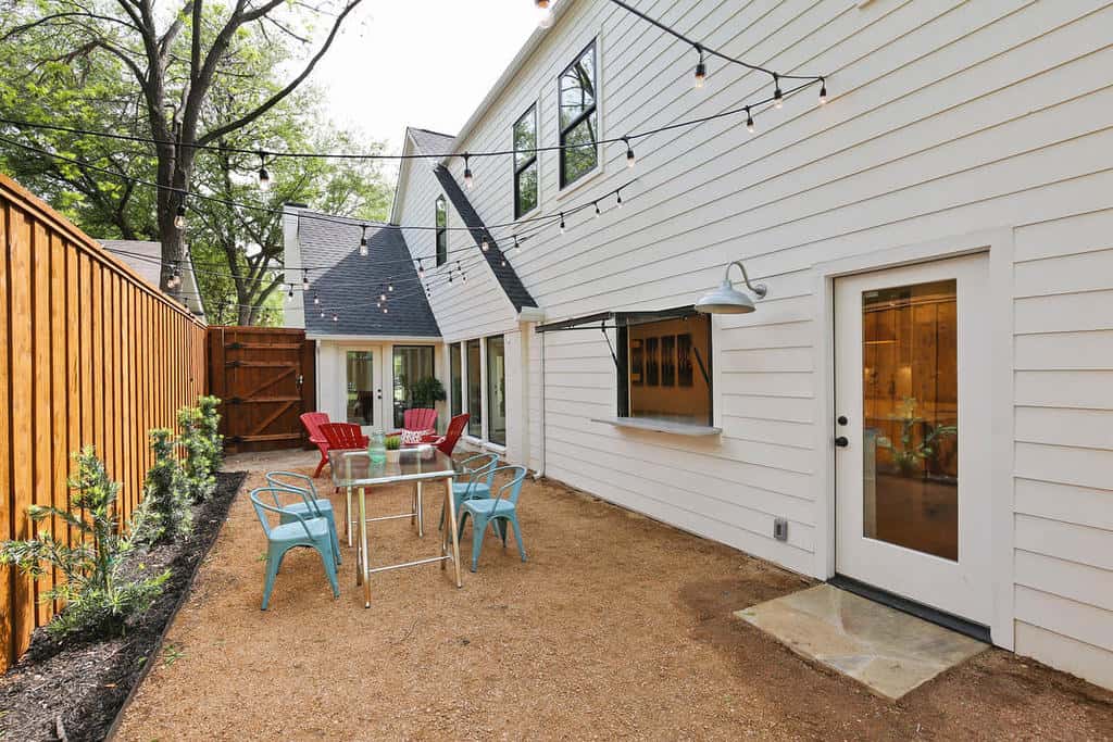 Bright backyard patio with string lights, red and teal chairs, a glass table, white house siding, and a wooden fence