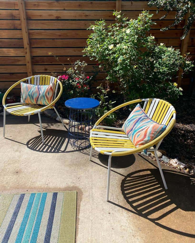 Two colorful chairs with striped cushions and a small blue table on a sunny patio, surrounded by plants and wooden fencing