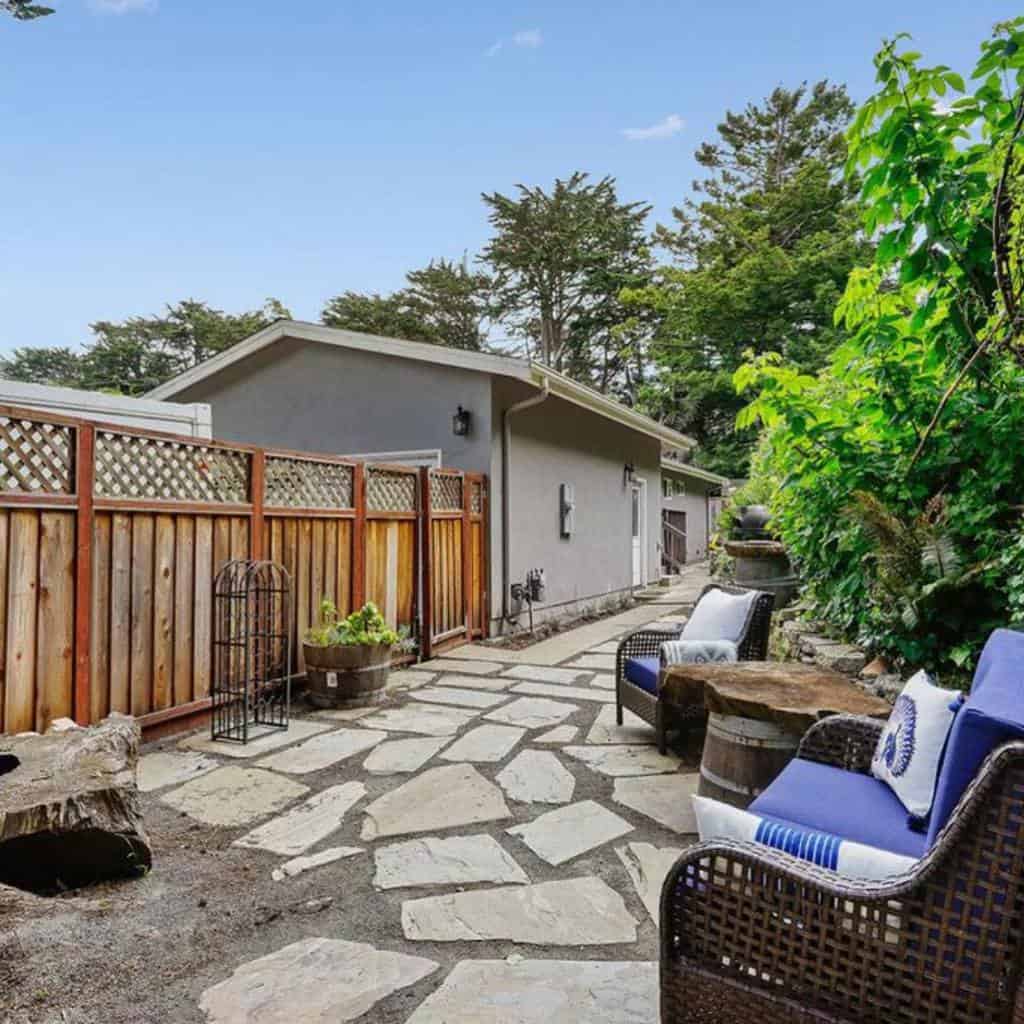 Side yard with a stone pathway, wooden fence, cozy wicker seating with blue cushions, lush greenery, and rustic barrel tables