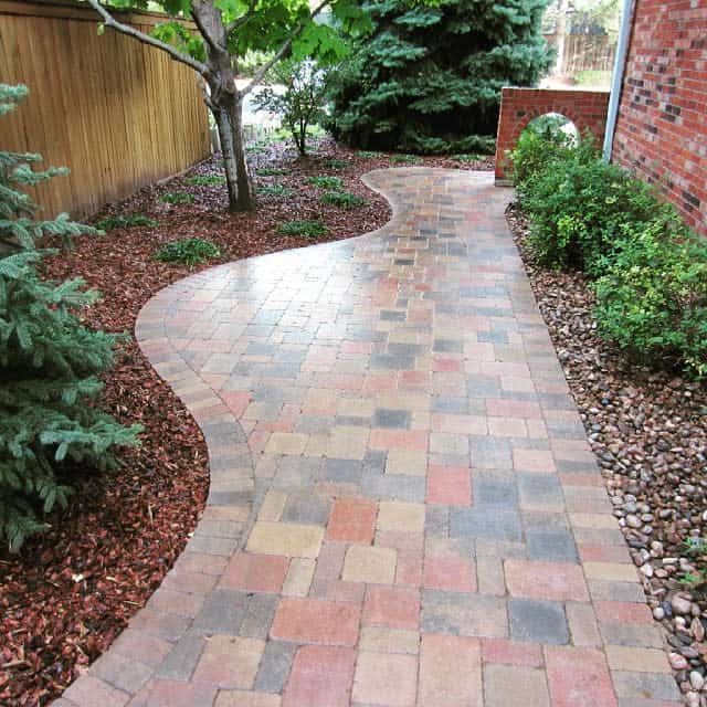 Curved brick pathway bordered by mulch and plants, leading past a wooden fence and red brick wall amidst greenery