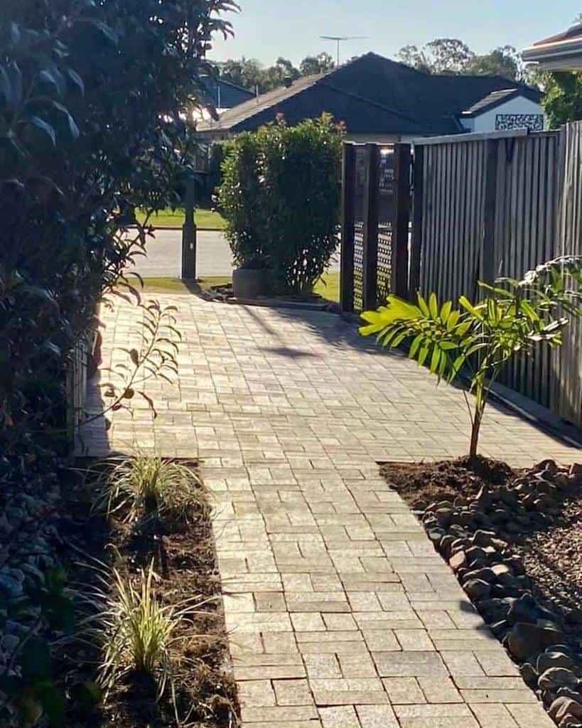 Brick pathway with small plants on the sides, leading to an open gate and a sunny street beyond