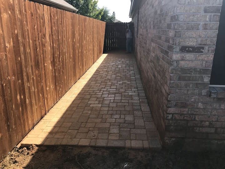 A narrow stone pathway between a wooden fence and a brick house, leading to a gated area