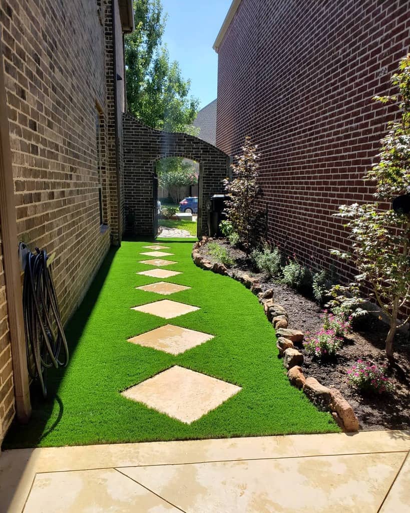 Narrow side yard with a green artificial lawn, stepping stones, and bordered by flower beds, leading to a sunny archway