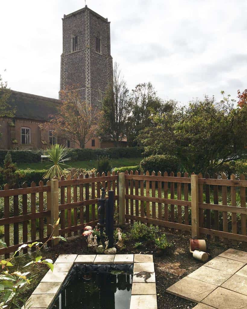 Charming wooden picket pallet fence surrounding a small garden pond, creating a peaceful outdoor space with a historic church view