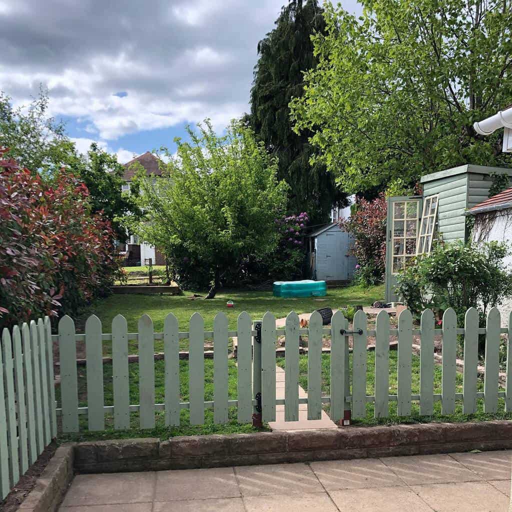 Charming light green picket fence with a small gate, enclosing a lush backyard garden with a cozy shed and mature trees