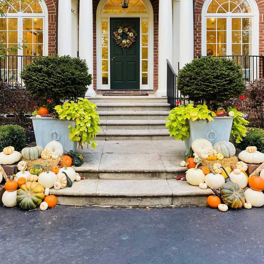 Front porch with pumpkins and gourds of various colors and sizes, two large potted shrubs, and a green door with a wreath