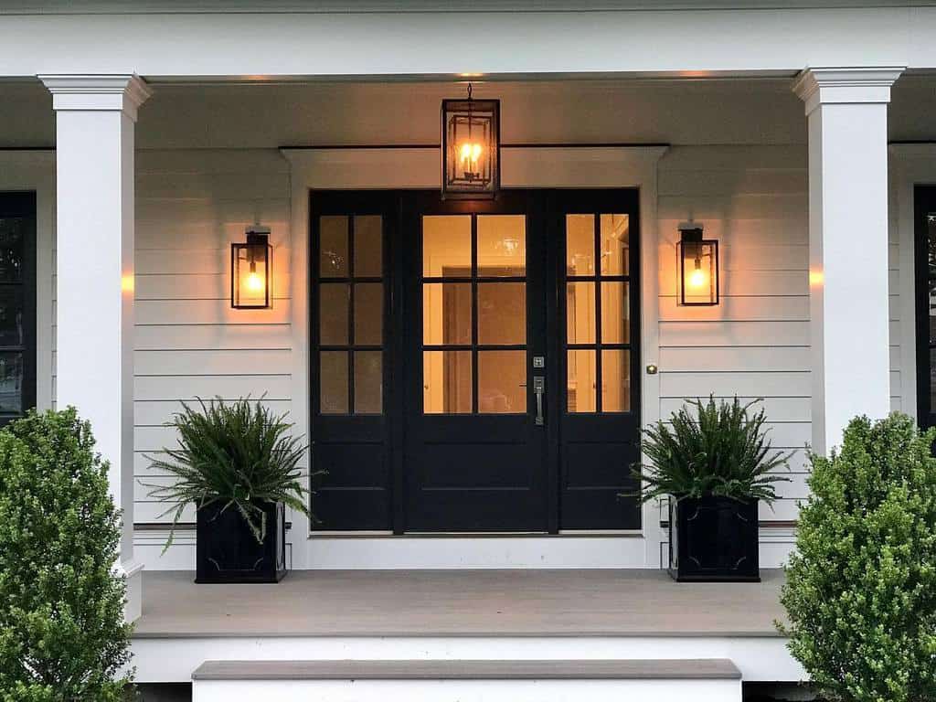 A front porch with a black door, lantern-style lights, and potted plants on each side, surrounded by white siding and columns framing the entrance