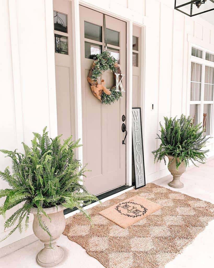 Elegant porch with a neutral front door, greenery-filled urns, a woven rug, and a welcoming wreath, creating a cozy and inviting entry