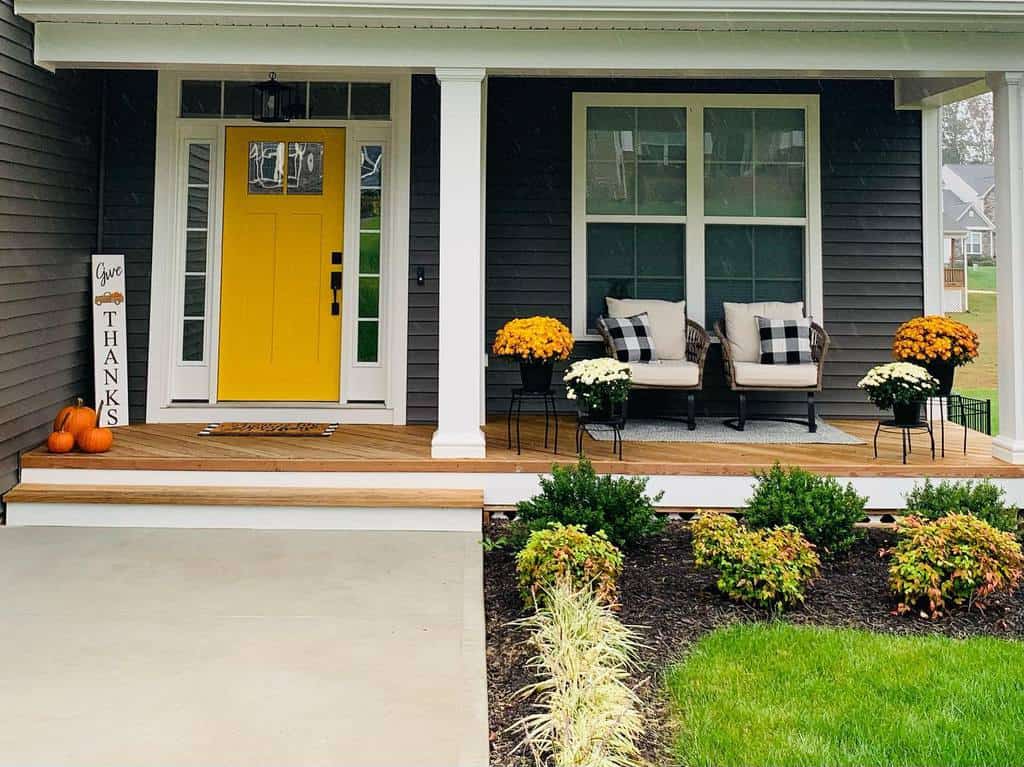 A welcoming porch with a bright yellow door, fall flowers, pumpkins, and two chairs with plaid cushions