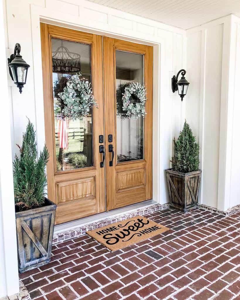 Wooden double doors with wreaths, flanked by potted plants. A doormat reads "Home Sweet Home" on a brick porch