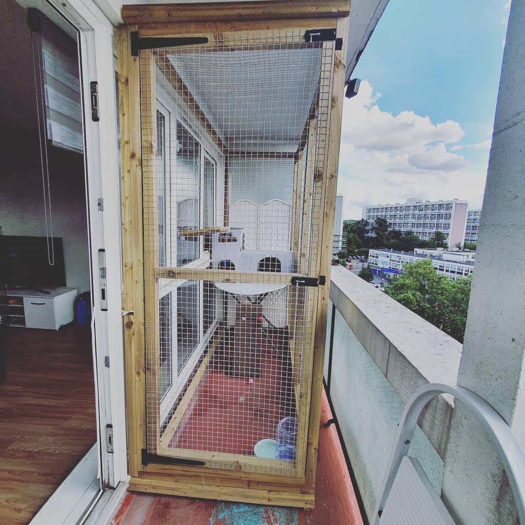 Enclosed balcony catio with a wooden frame and mesh walls, providing a secure outdoor space with climbing shelves and cozy seating for cats