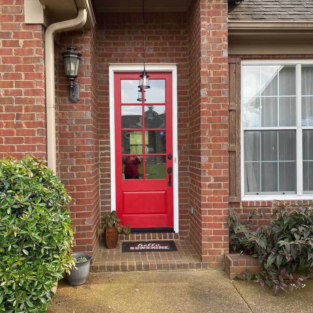 A red front door with a hanging lantern above, surrounded by brick walls, plants on each side, and a doormat in front