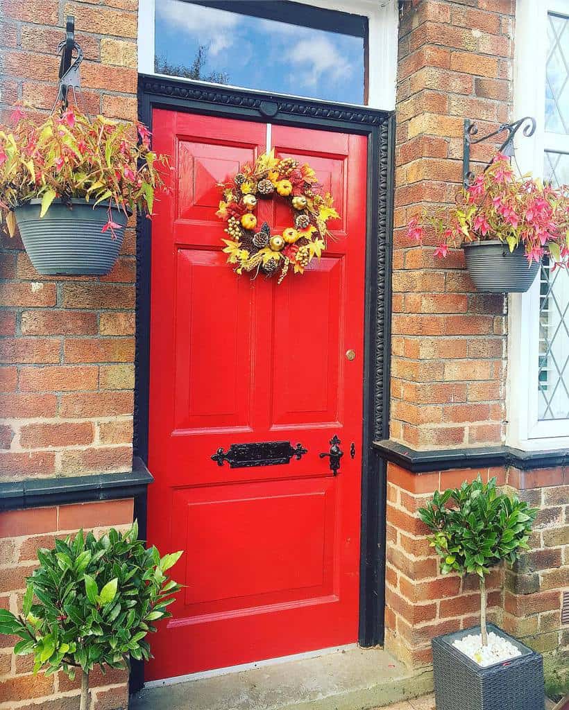 A bright red door with a fall wreath, flanked by hanging plants and potted shrubs, set in a brick wall with a window on the right