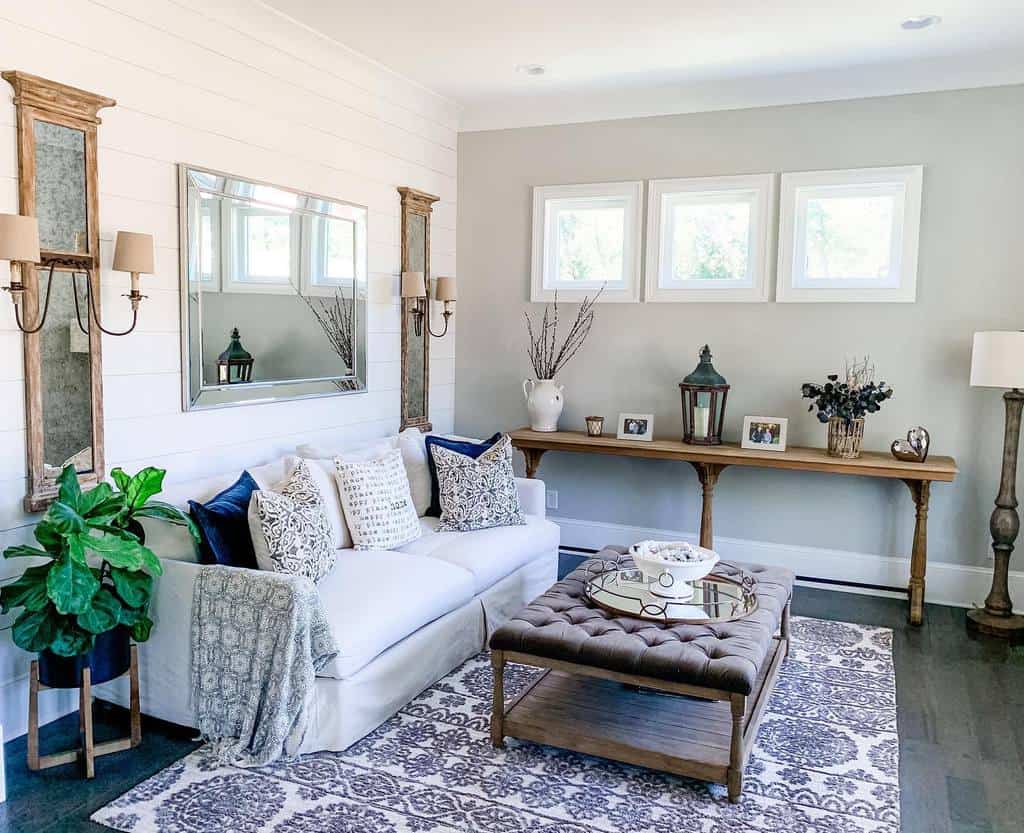 A cozy living room with a white sofa, decorative pillows, a tufted ottoman, and a wooden console table with plants and framed photos