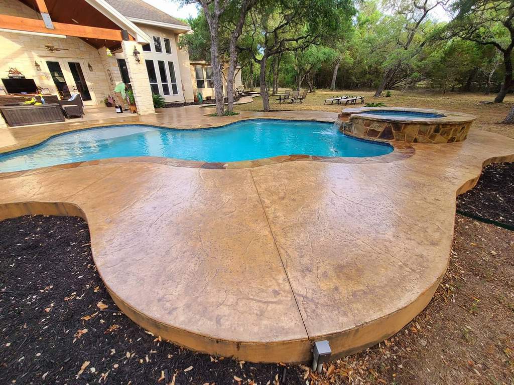 Backyard view of a house with a uniquely shaped swimming pool and hot tub, surrounded by trees and a stone patio