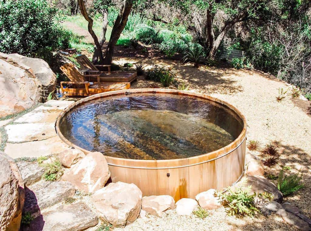 A round wooden hot tub filled with water is surrounded by rocks, deck chairs, and trees in a sunny garden setting