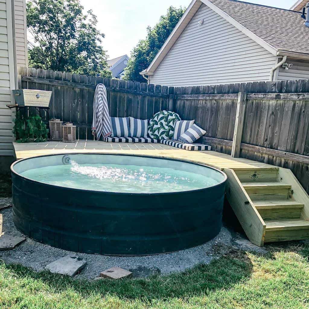 Outdoor stock tank pool surrounded by a wooden deck, with a striped lounge chair and greenery next to a wooden fence