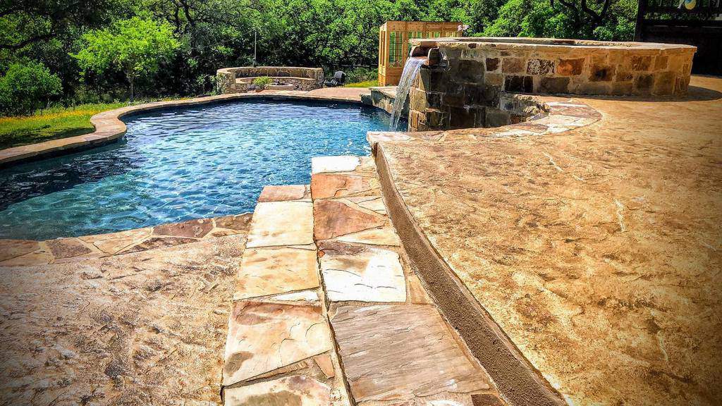 A stone patio with a curved pool featuring a waterfall, surrounded by trees in the background