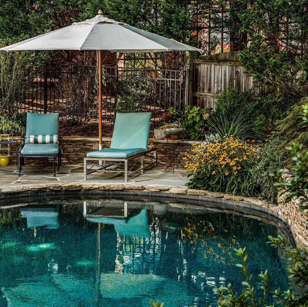 Poolside scene with two lounge chairs under a white umbrella, surrounded by greenery and flowers, patio area with stone tiles