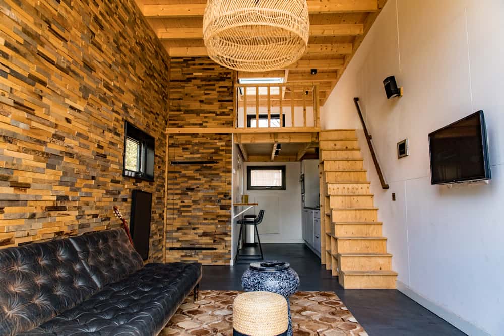 Rustic tiny home with a lofted bedroom, wooden staircase storage, and textured wood walls. A black leather couch adds contrast to the space.