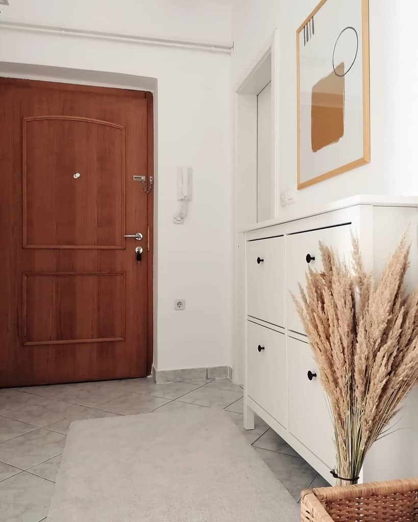 Modern minimalist entryway with a sleek white shoe cabinet, warm wood door, abstract artwork, and natural pampas grass decor