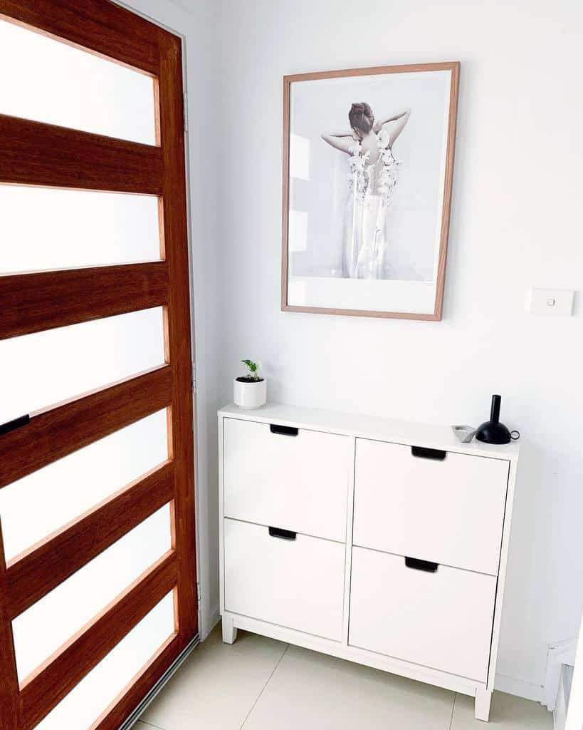 Minimalist entryway with a sleek white shoe cabinet, warm wooden door, black-and-white artwork, and simple modern decor