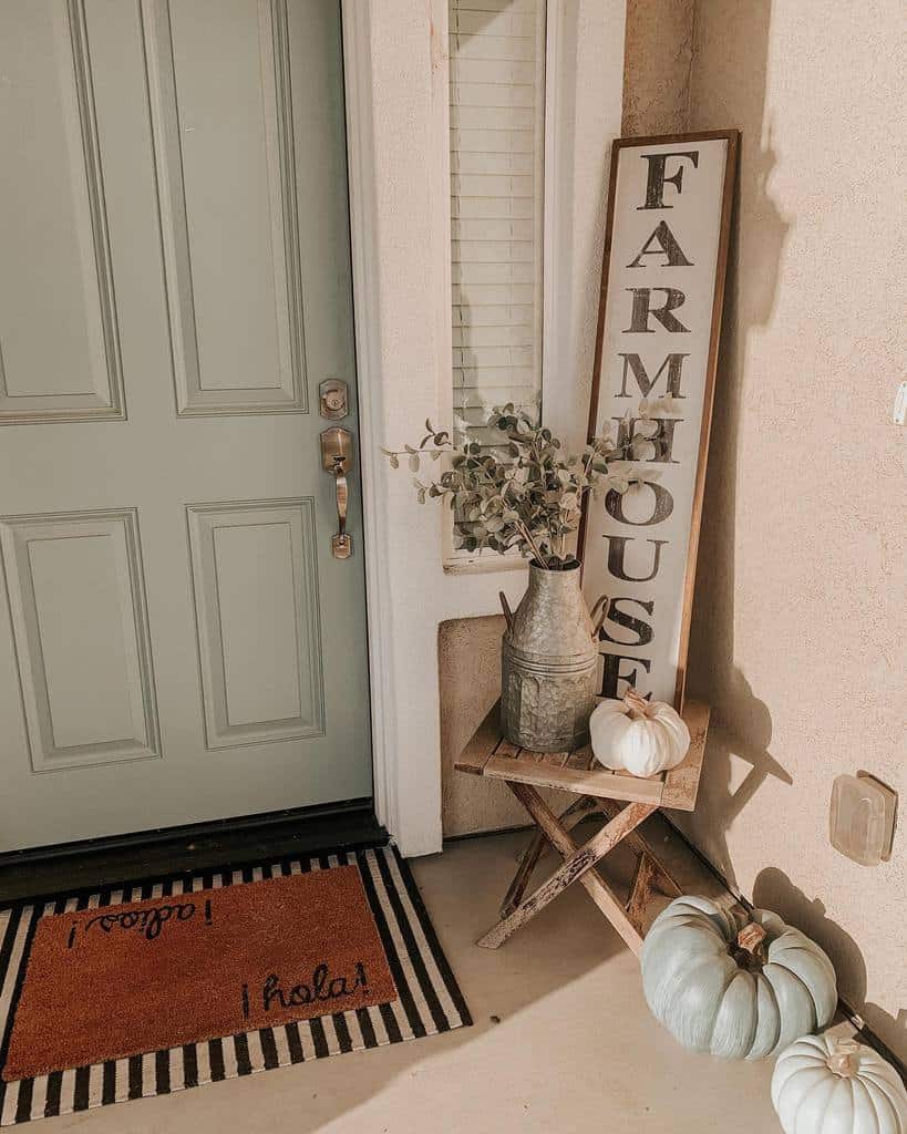 Cozy farmhouse-style porch with a muted green door, rustic decor, a "Farmhouse" sign, and pastel pumpkins for a warm, inviting entryway