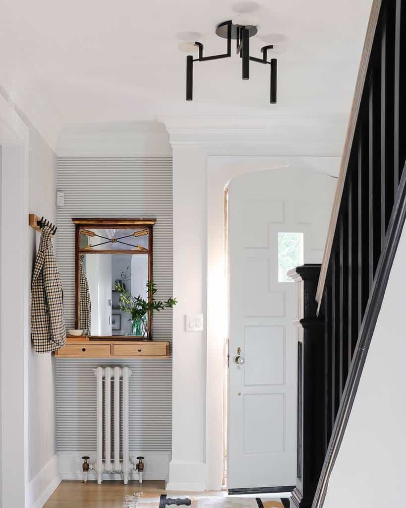 Chic entryway with a vintage wall-mounted console, classic mirror, striped wallpaper, modern lighting, and a stylish black staircase