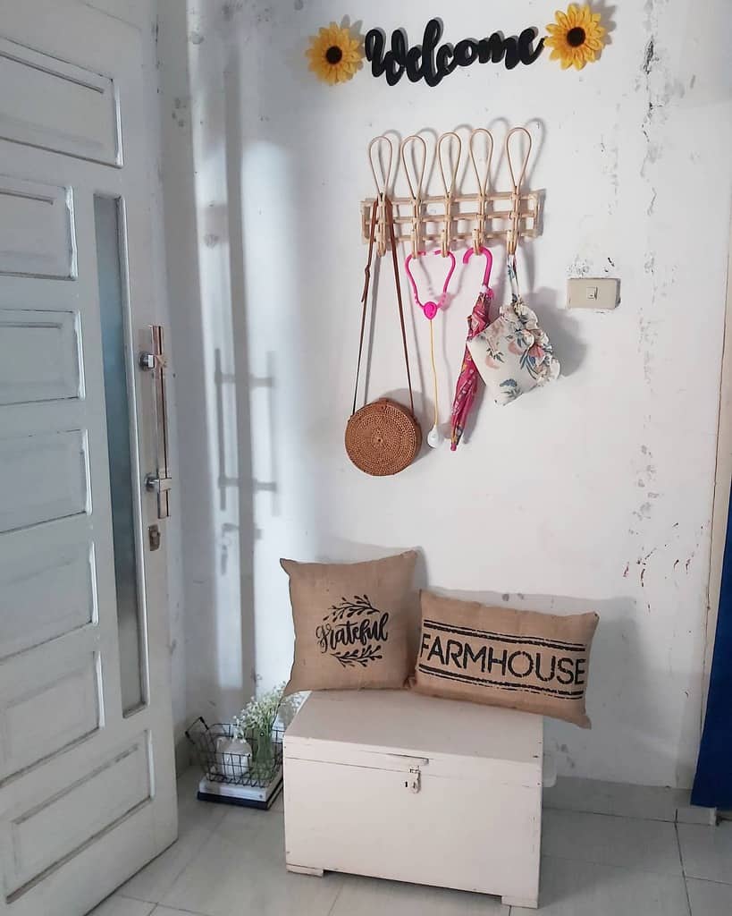 Cozy farmhouse-style entryway with a white storage bench, burlap pillows, woven wall hooks, and a welcoming sunflower decor accent